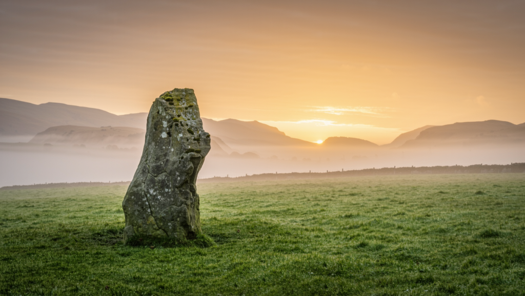 A single weathered standing stone in a quiet grass field at sunrise with mist over distant hills